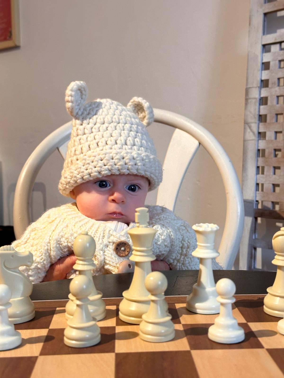 A baby in a cream jumper watching a chess set at the club